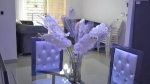 Un comedor moderno con mesas y sillas de vidrio, un jarrón central con flores blancas e iluminación colgante.