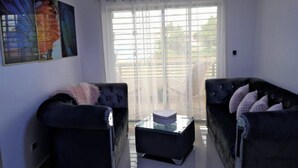 Una sala de estar con dos sofás negros, una mesa de centro de cristal y un ventanal con persianas.
