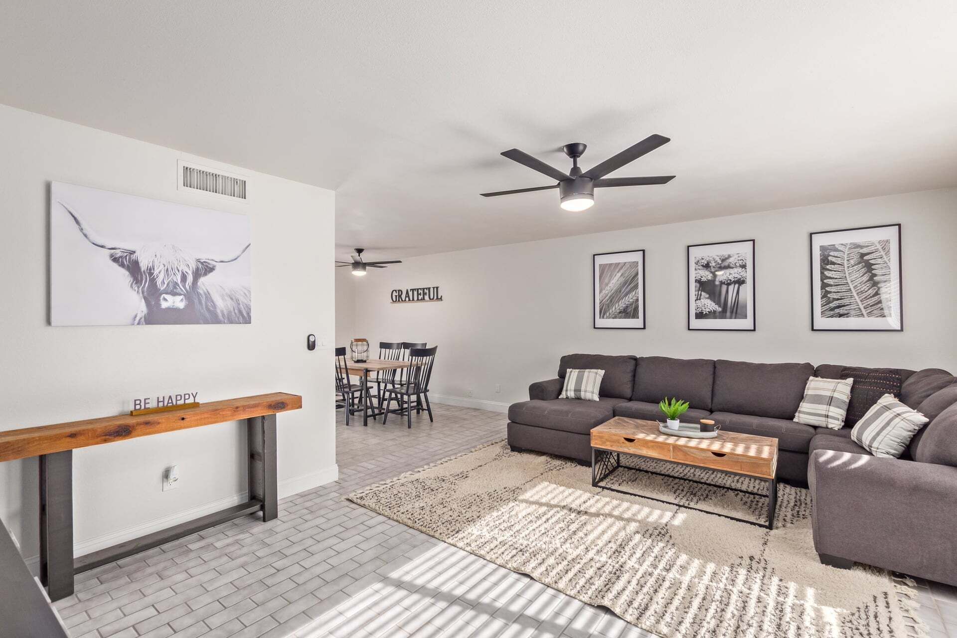 Een moderne woonkamer met een grijze bank, een houten salontafel en een eethoek met een witte tafel en stoelen.