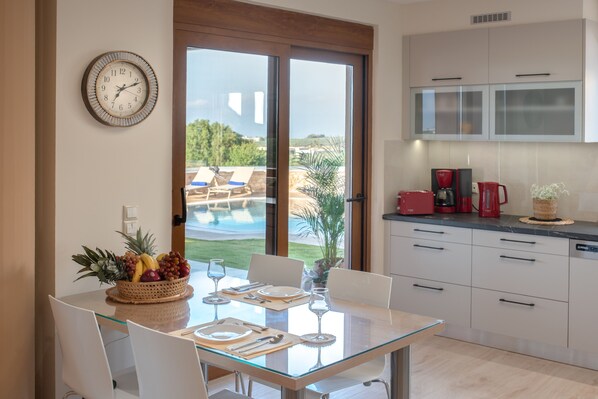 Una cocina moderna con una mesa de comedor de vidrio, armarios blancos y vistas a una piscina y vegetación al exterior.