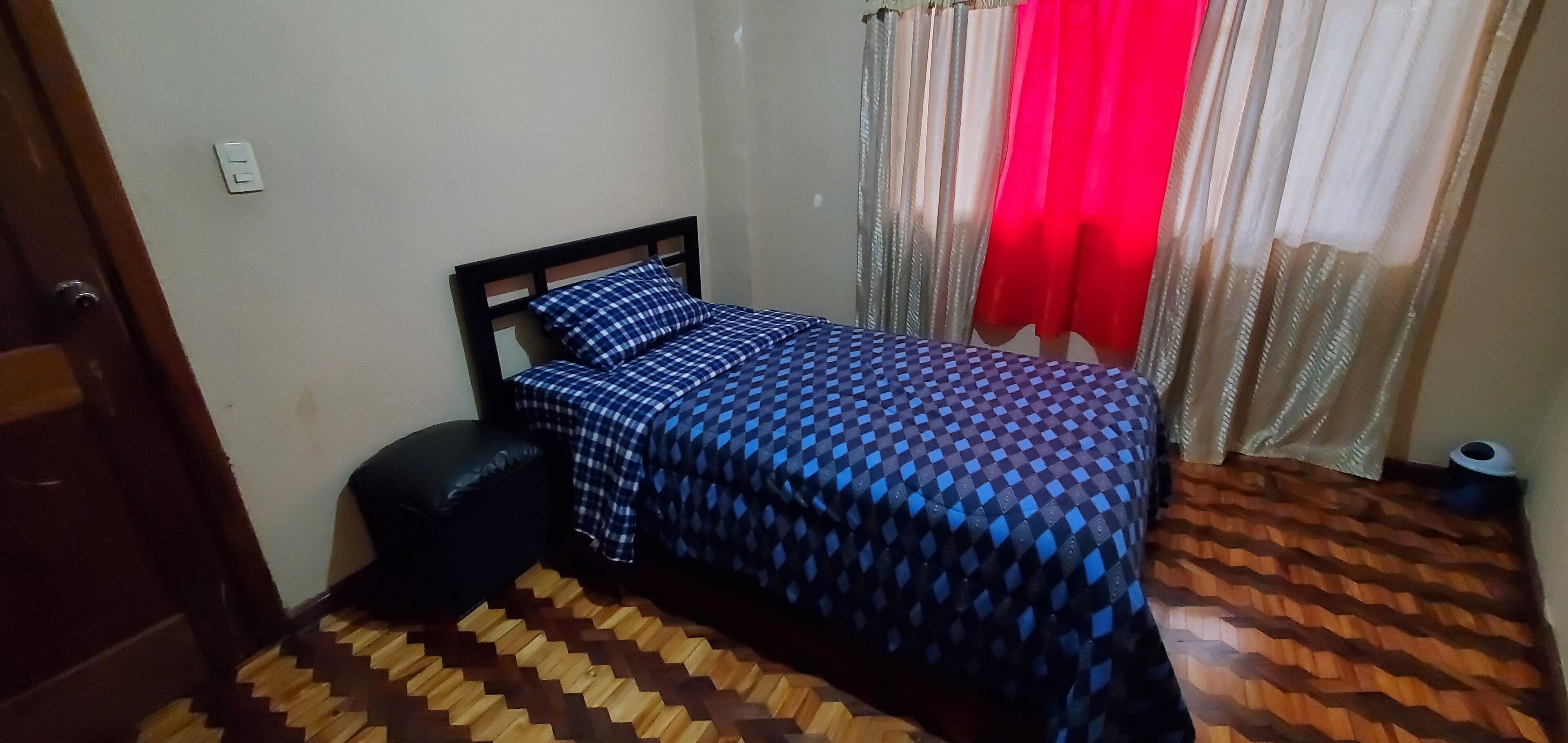 A bedroom with a bed, a small stool, a wooden floor, and a door.