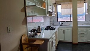 A kitchen with white cabinets, a wooden chair, and a view of a town through the window. - Apartamento 3 habitaciones y parqueo (Cuenca)