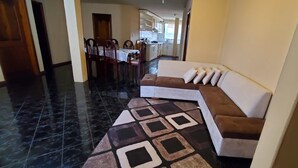 A modern living room with a sectional sofa, a dining area with a table and chairs, and a kitchen in the background. - Apartamento 3 habitaciones y parqueo (Cuenca)