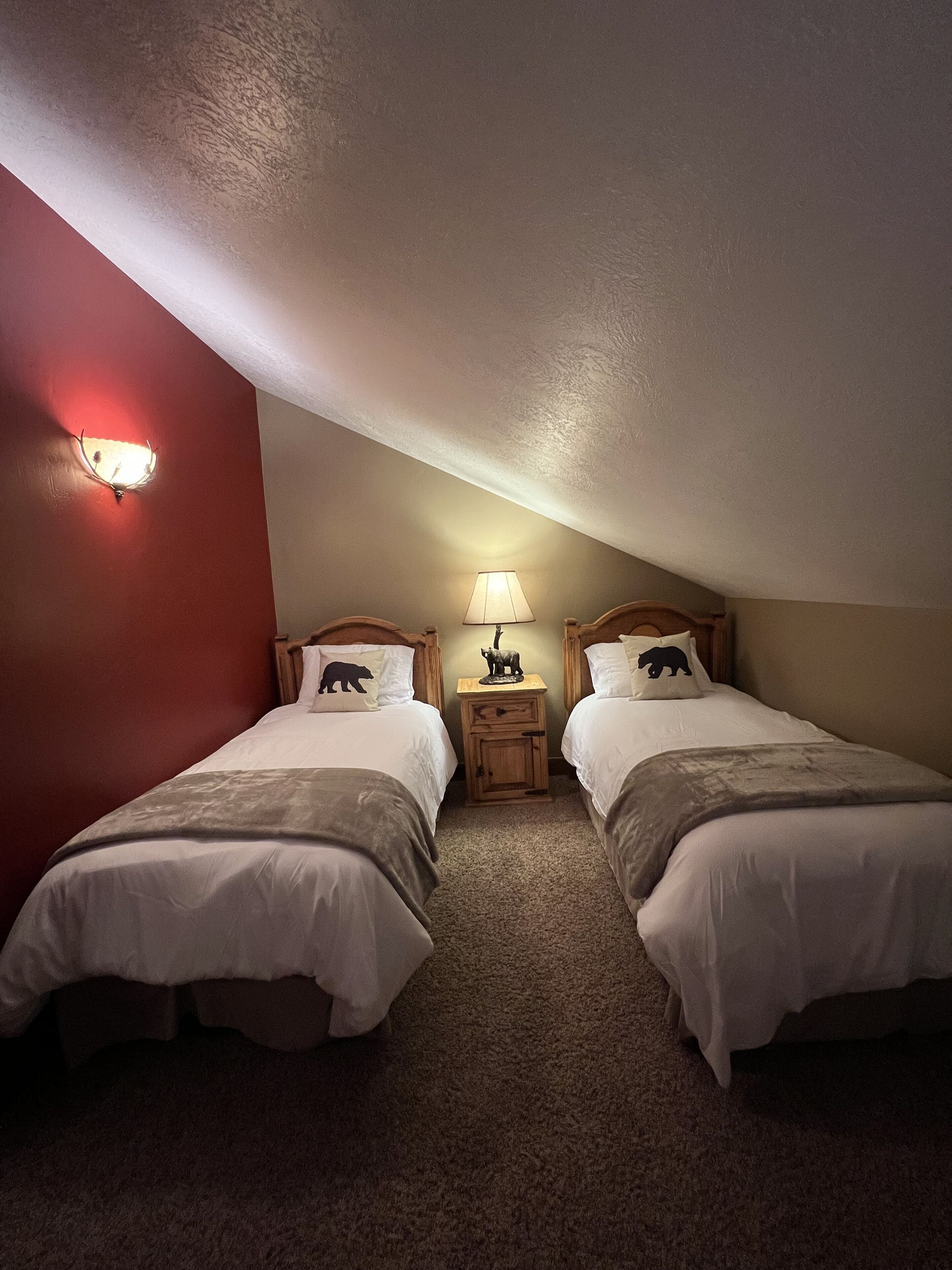 Une chambre avec deux lits, un mur d’accent rouge, une petite table de chevet en bois avec une lampe, et un plafond à la finition texturée.