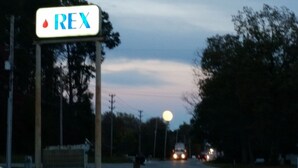 Een bord van een Rex benzinestation, met een heldere lucht en een volle maan op de achtergrond.