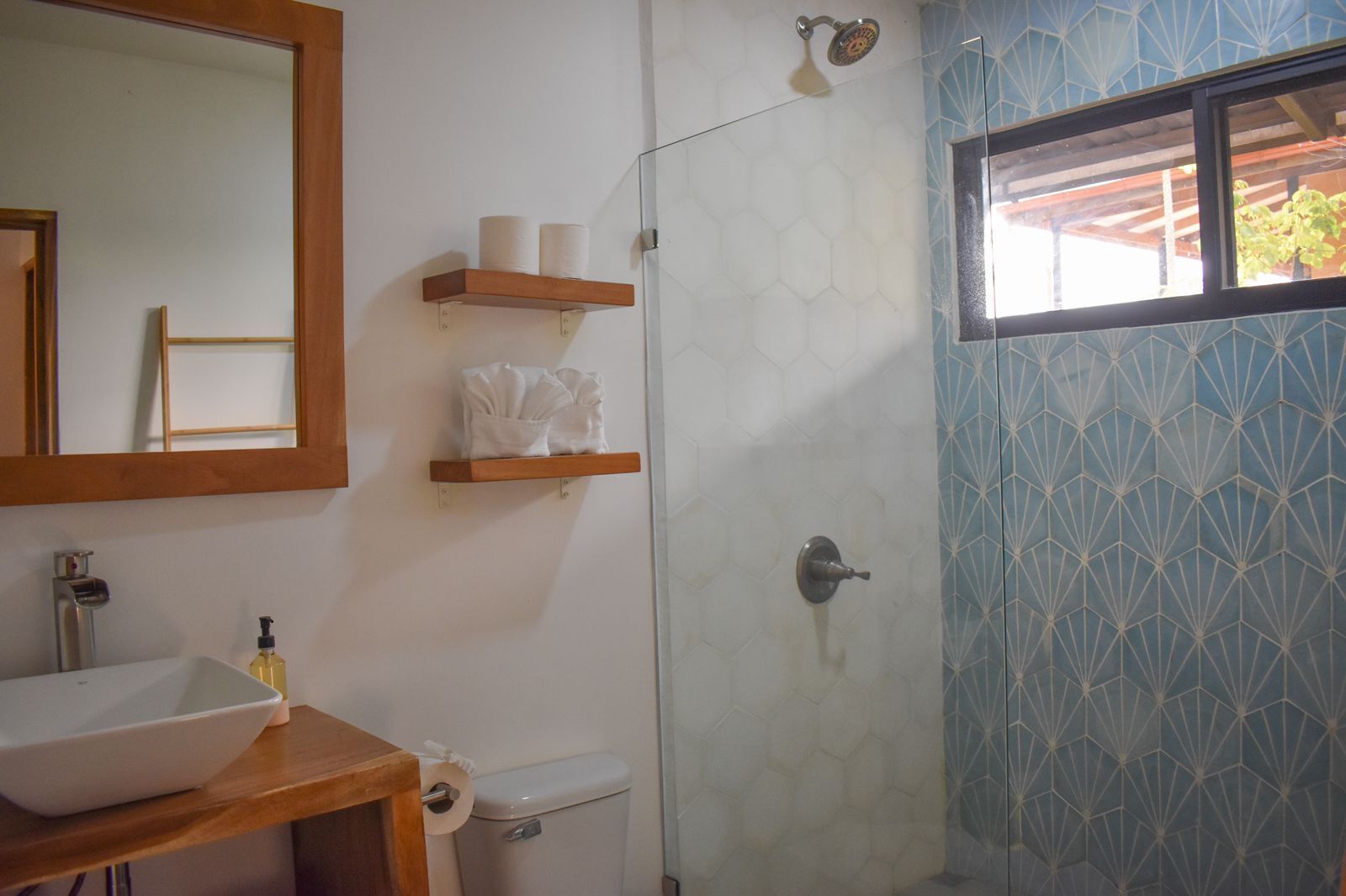 Un baño con tocador de madera, inodoro, ducha con puerta de vidrio y ventana con diseño geométrico.