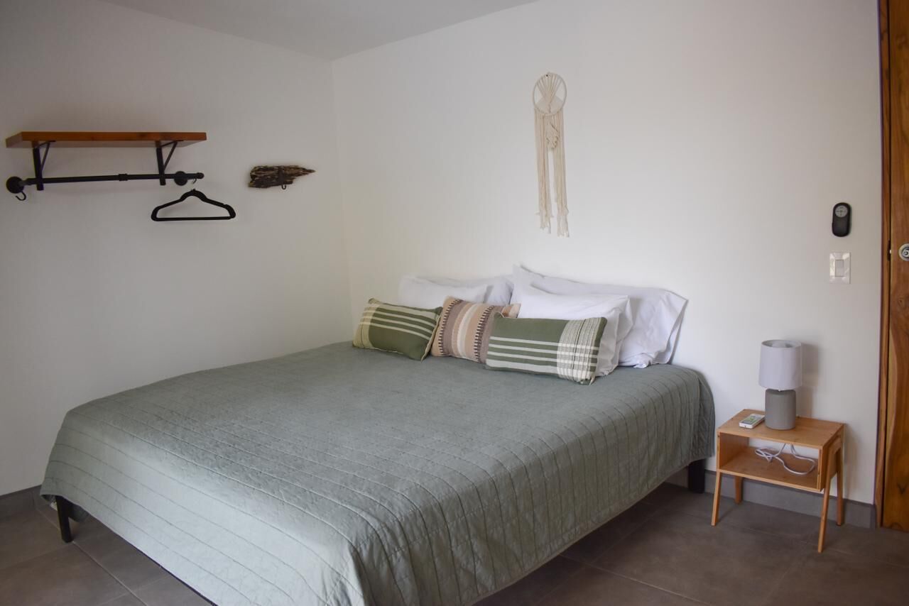 Un dormitorio con cama, mesita de noche con lámpara, perchero y un atrapasueños colgado en la pared.
