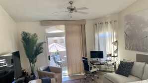 A living room with a ceiling fan, a TV, a desk with a computer, and a sofa. There is a large window with curtains and a view of the outside. - Paradise Valley area condo with heated pool and gym,  near Mayo Clinic. (Scottsdale)