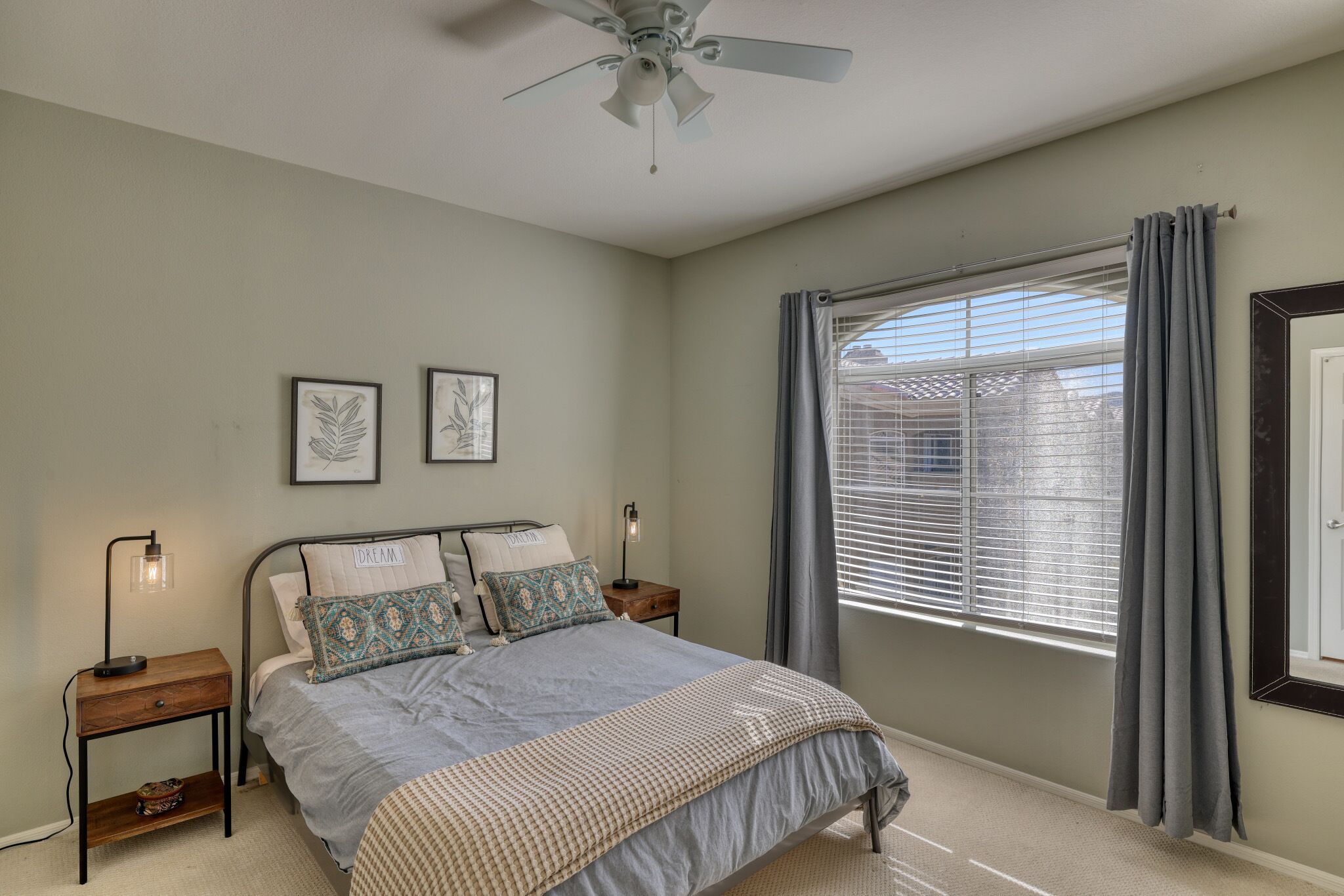 A bedroom with a bed, nightstands, a ceiling fan, a window with curtains, and framed artwork on the wall.