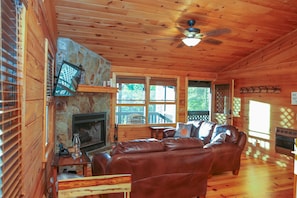 A cozy cabin interior with a stone fireplace, leather sofa, and large windows.