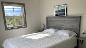 Un lit bien fait, avec une tête de lit, une fenêtre donnant sur les arbres et le ciel, et un tableau encadré accroché au mur.