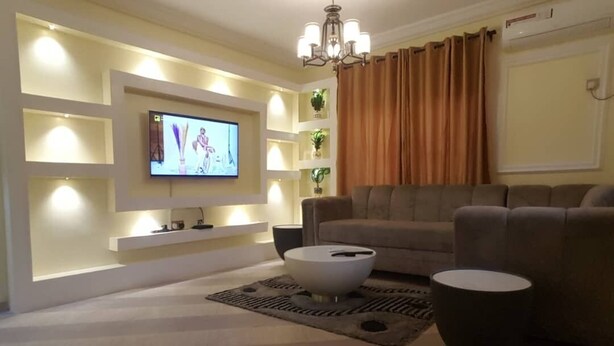 A living room with a flat-screen TV mounted on a wall, a sofa, two black side tables, and a patterned rug. - Have a Quality time with your Family in this  3bed apartment on the ground floor (Lagos)