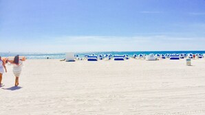A beach with rows of blue and white beach umbrellas and lounge chairs, a sandy beach, and the ocean in the background.