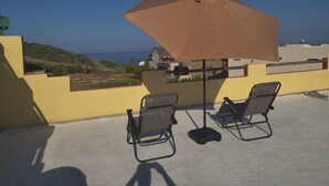 Een balkon met twee stoelen en een grote parasol, met uitzicht op een kustlandschap met gebouwen en begroeiing.