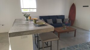 Een ruimtelijk ingerichte leefruimte met een kitchenette, een bank, een surfplank en een klein bijzettafeltje.