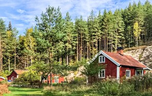 Une maison en bois rouge, avec un toit en pente, entourée d’une zone boisée où poussent de grands pins.