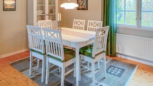 Zona pranzo con tavolo e sedie bianche, lampadario a sospensione, mobile con ante in vetro e finestra con tende.