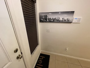 Un pasillo con una puerta, una ventana con persianas, un termostato en la pared y un felpudo con la palabra 'hello'.