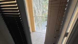 Vista desde un balcón con baranda metálica, piso de concreto y puerta que da a un área exterior con árboles y una cerca.