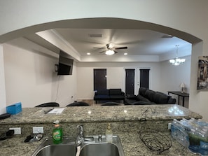 Una cocina con encimeras de granito, un lavamanos y un ventilador de techo. Hay una sala de estar con un sofá y una televisión montada en la pared.