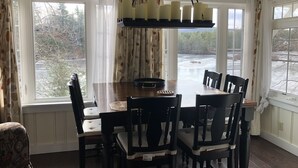 Een eetgedeelte met een houten tafel en stoelen, grote ramen met gordijnen, en een hanglamp.