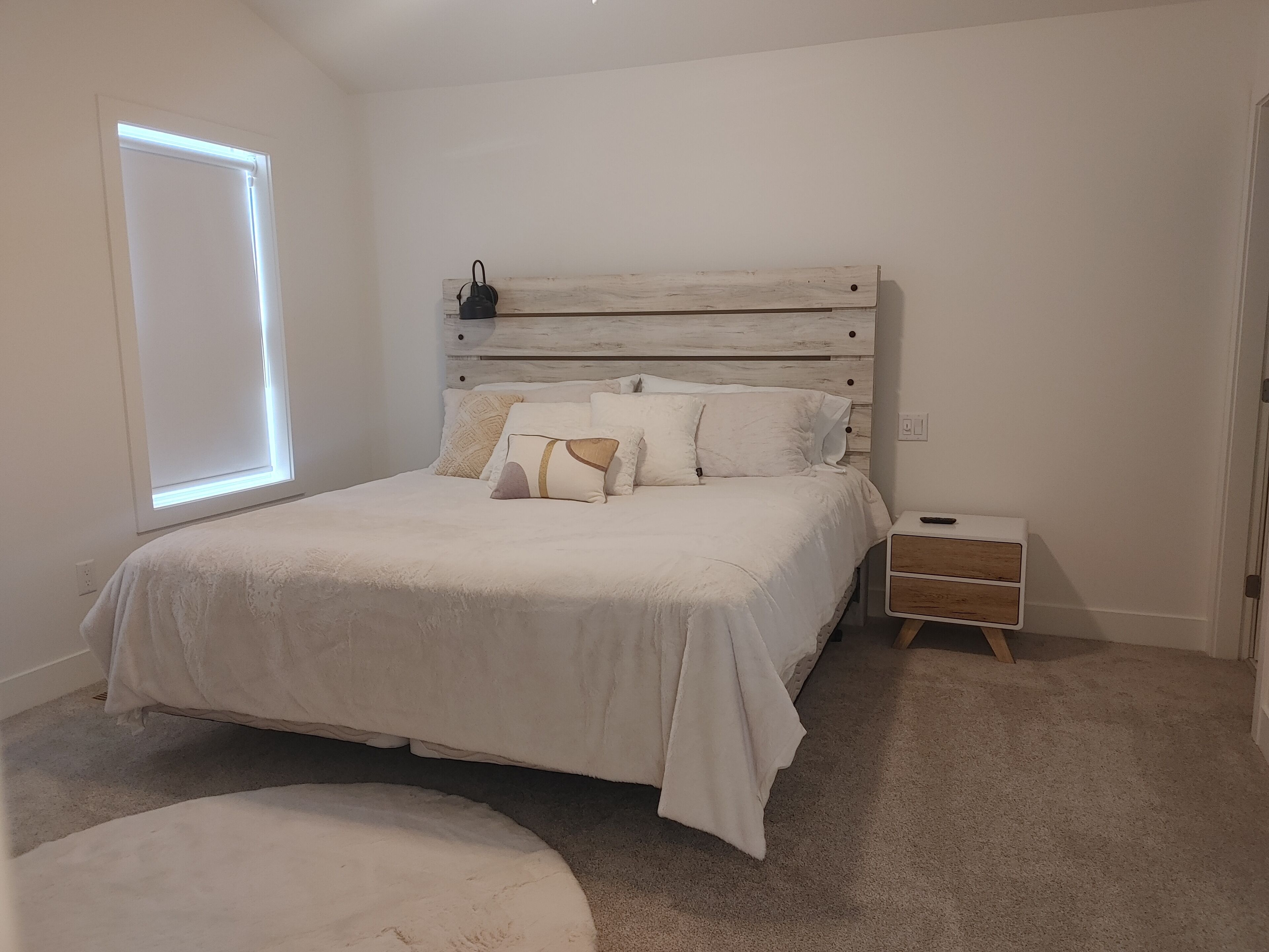 A bedroom with a bed, a bedside table, a nightstand, a window, and a carpeted floor.