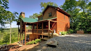 A log cabin with a porch, stone chimney, and a view of the surrounding forest.