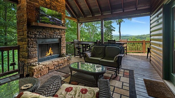 A covered outdoor area with a stone fireplace, seating, and a view of the surrounding forest.