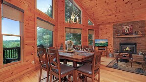 A wooden cabin interior with a dining area, fireplace, and large windows overlooking a forest.