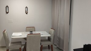 Uma área de refeições com uma mesa retangular, quatro cadeiras, dois espelhos fixados na parede, uma luminária no teto, uma janela com cortinas e uma pequena mesa com um objeto preto.