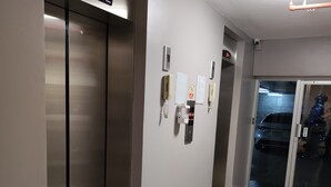 Um elevador com mostrador digital, interfone na parede e porta de vidro, onde se vê um carro do lado de fora.