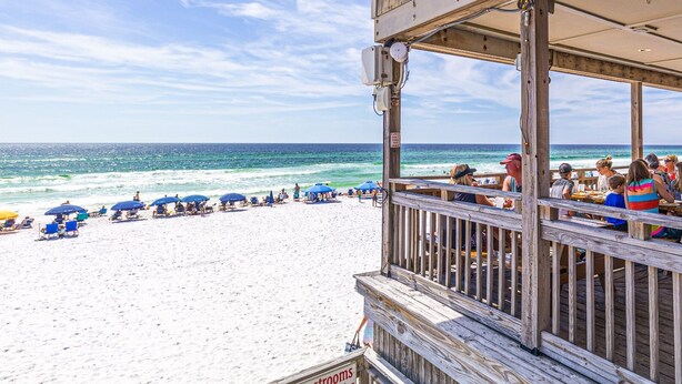 A beachside restaurant with a wooden deck overlooking the ocean, featuring outdoor seating and umbrellas.