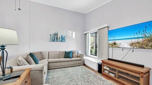 A living room with a beige sofa, a wooden console table, a large flat-screen TV, and a painting of a beach scene.