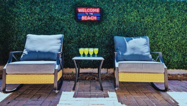 Dos sillones con cojines, una mesita con bebidas y un cartel que dice 'Welcome to the Beach' frente a un seto verde.