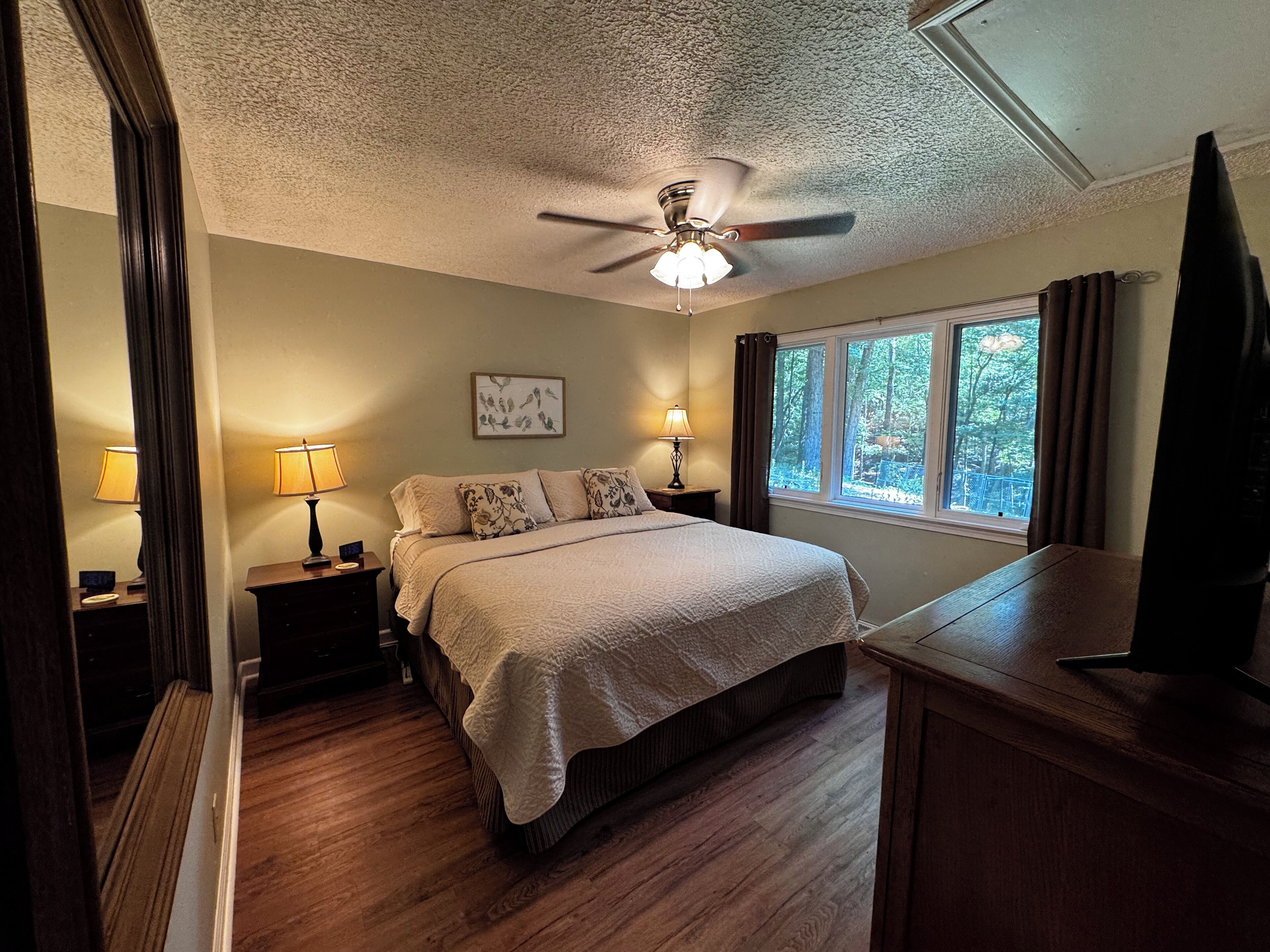 Une chambre à coucher avec un lit, deux tables de chevet avec des lampes, un ventilateur de plafond, une fenêtre avec des rideaux et un téléviseur posé sur un meuble.