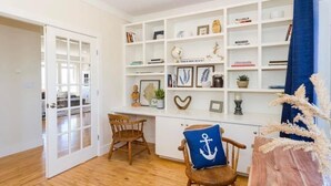 Ein Zimmer mit Holzboden, einem weißen Regal mit verschiedenen Dekorationen, einem blauen Kissen mit Anker-Motiv und einer Tür zu einem weiteren Raum.