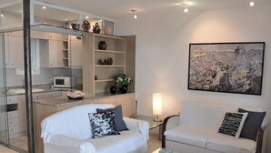 A modern living room with a glass coffee table, two white sofas with black and white cushions, a kitchen with a microwave, and a cityscape painting on the wall. - Apartament Bloc Goya (Begur)