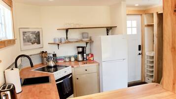 Una cucina compatta con un piano di lavoro in legno, un fornello, un lavandino e un frigorifero bianco. Sui pensili sopra il bancone si trovano piatti e bicchieri.