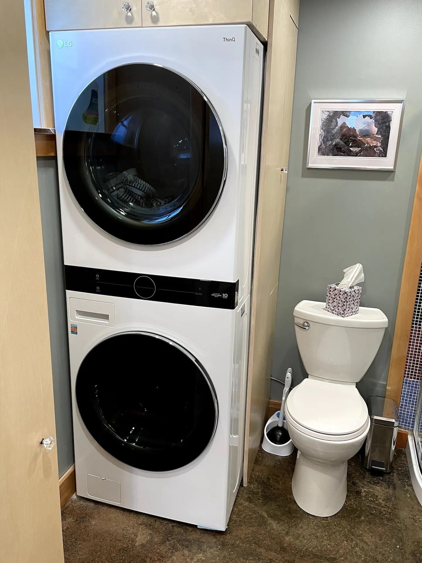 A compact washer and dryer unit in a bathroom, with a toilet and a framed picture on the wall.