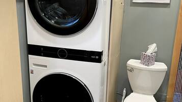 A compact washer and dryer unit in a bathroom, with a toilet and a framed picture on the wall.