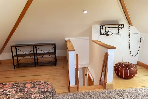 A loft bedroom with a wooden floor, a small wooden table, a hanging light fixture, and a decorative pouf.