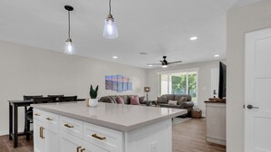 Une cuisine moderne avec des meubles blancs, un îlot central, des luminaires suspendus et un coin repas avec une table et des chaises aux couleurs sombres.