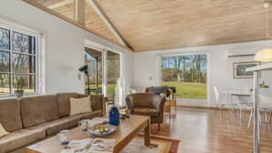 A spacious living area with a wooden ceiling, large windows offering views of a grassy yard, a sofa, a coffee table with reading materials, and a dining area with chairs and a table.