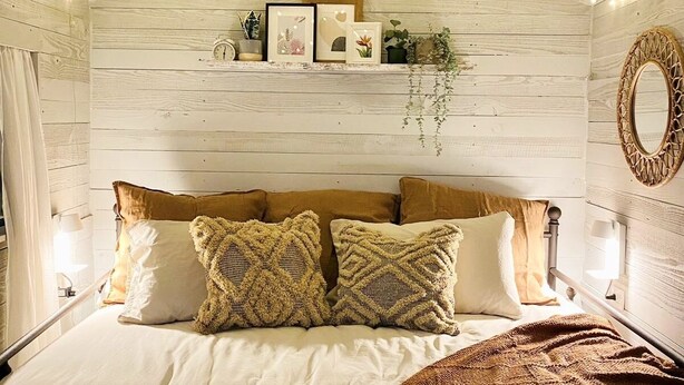 Une chambre avec un plafond en bois, des guirlandes lumineuses, un lit agrémenté d’oreillers à motifs, une table de chevet avec une lampe et un miroir fixé au mur.