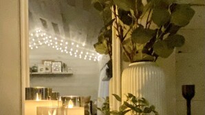 Une pièce avec un miroir qui reflète une guirlande lumineuse, un vase blanc contenant une plante et deux bougies allumées sur une table.