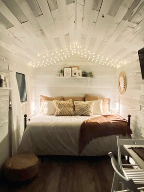 A cozy bedroom with a wooden ceiling, a bed with patterned pillows, a bedside table, and a round stool. - *Gorgeous tiny home in the city! (Miami)