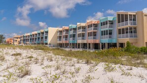A row of multi-story beachside buildings with balconies, situated on a sandy beach with sparse vegetation. - Direct beach access! Summer and fall dates still available, call today! (Bradenton Beach)