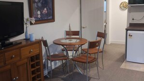 A compact living space with a dining table, chairs, a television, a refrigerator, and a door leading to another room.