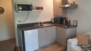 A compact kitchen with a dishwasher, microwave, and a small sink. There is a framed picture on the wall and a couch with a blanket in the adjacent living area.