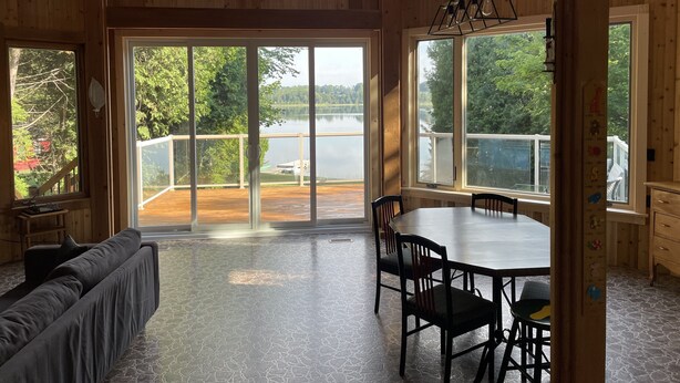Ein geräumiges Zimmer mit einem großen Schiebeglastür, die auf eine Terrasse mit Seeblick führt, Holzvertäfelung und einem Essbereich mit Stühlen und Tisch.